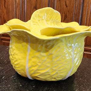 May include: A large, yellow ceramic cabbage-shaped bowl with a textured surface and white accents. The bowl has a lid that resembles cabbage leaves. The bowl is sitting on a dark surface.