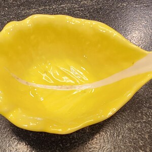 May include: A bright yellow, leaf-shaped ceramic dish with a white accent. The dish has a glossy finish and a curved, organic form. The white accent runs along the center of the dish, adding contrast. This decorative dish is suitable for serving small portions.