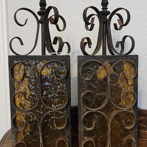 May include: Pair of vintage wrought iron sconces. Each sconce features a rectangular frame with textured amber glass and decorative scrollwork. The sconces have a dark brown finish and a pointed finial at the top.