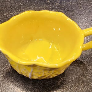 May include: A bright yellow ceramic gravy boat with a handle. The boat has a scalloped edge and a textured, possibly woven, design on the lower portion. The interior is smooth and glossy.