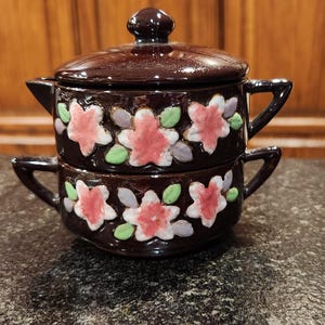 May include: A dark brown ceramic soup tureen with a lid, featuring a floral design with pink, white, and green accents. The tureen has two handles and is stacked on a matching bowl, creating a charming, vintage aesthetic.