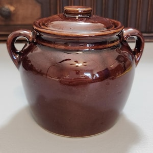 May include: A small, brown ceramic jar with a lid and two handles. The jar has a glossy finish and a crack on the side. The lid has a small knob on top. The jar is likely used for storage.