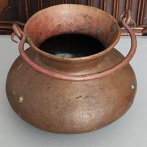 May include: A copper pot with a rounded body and a wide, open top. The pot has a curved handle and a dark interior. The copper has a weathered appearance with some green and brown spots, suggesting age and use. The pot is likely used for cooking or storage.