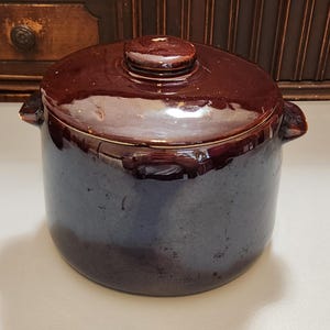 May include: A dark brown ceramic pot with a matching lid. The pot has two small handles on either side and a round knob on the lid. The pot is glazed and has a glossy finish. The pot is likely used for cooking or storage.