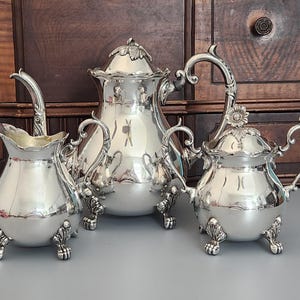 May include: A silver tea set including a teapot, creamer, and sugar bowl. The teapot has a lid with a leaf-shaped finial. The creamer and sugar bowl have ornate handles and are supported by decorative feet. The set is displayed on a gray surface against a dark wood background.