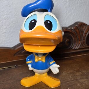 May include: A Donald Duck bobblehead figurine. The figurine features a blue sailor hat, a white face with large blue eyes, and an orange beak. Donald wears a blue sailor shirt with a yellow bow tie and yellow feet. The figurine is a collectible item.