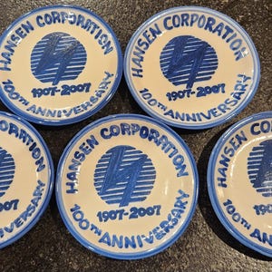 May include: Five white ceramic plates with blue rims and text. Each plate features the words "Hansen Corporation" and "100th Anniversary 1907-2007" in blue. A blue graphic is in the center.
