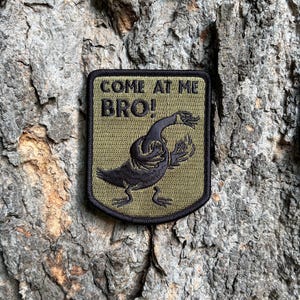 May include: A rectangular olive green patch with a black embroidered border. The patch features a black embroidered graphic of a goose with its wings raised in a fighting stance and the black embroidered text "COME AT ME BRO!" above it.