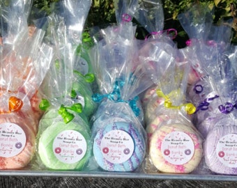 May include: A tray of colorful donut-shaped bath bombs, each individually wrapped in clear cellophane and tied with a ribbon. The bath bombs are in various colors, including pink, green, blue, and purple, with a label that reads "The Beauty Bar Soap Co. Donut Bath Bombs".