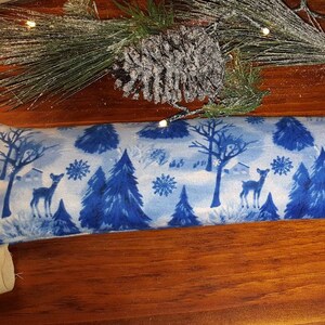 May include: A blue and white fabric rectangular pillow with a winter scene design. The design features trees, snowflakes, and deer.
