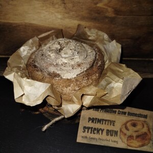 May include: A brown cinnamon roll with a white icing drizzle, wrapped in brown paper and tied with twine. The roll is labeled "Primitive Sticky Bun with Gooey Drizzled Icing" and is from "The Primitive Crow Homestead."