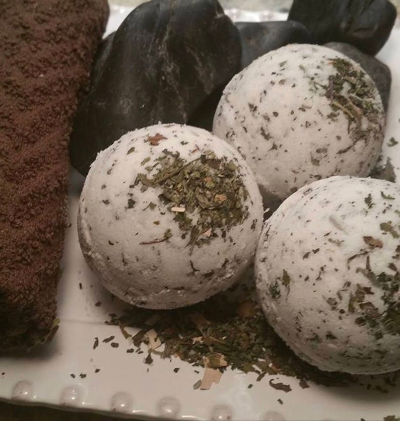 May include: Three white bath bombs with green herbs on a white surface. The bath bombs are round and have a rough texture.