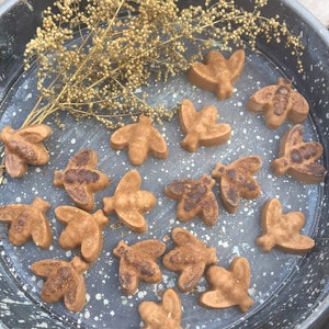 May include: A collection of 16 brown bee-shaped soaps in a metal bowl. The soaps are arranged in a random pattern and are surrounded by dried flowers.