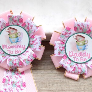 May include: Two pink and white teacup themed baby shower sashes with gold trim. One sash says "Mommy To Be" and the other says "Daddy To Be".