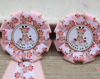 May include: Two pink and white ribbon rosettes with gold trim. The rosettes feature a giraffe illustration and the text "Mommy to be" and "Daddy to be".
