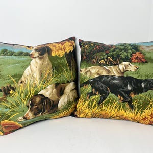 May include: Two decorative throw pillows with dog illustrations. One pillow features a brown and white dog sitting in a field. The other pillow has two dogs in a field, one black and one tan.