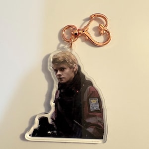 May include: A clear acrylic keychain featuring a portrait of a person with blonde hair, wearing a dark jacket with a red and black patch. The keychain has a rose gold heart-shaped clasp.