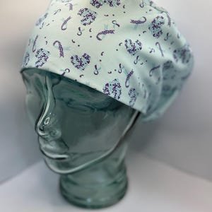 May include: A light blue surgical scrub cap with a repeating pattern of purple awareness ribbons and heart shapes. The cap is made of a soft fabric and is designed to cover the hair during medical procedures.