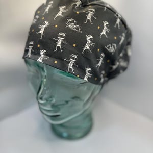 May include: A black scrub hat with a white mummy pattern. The hat has a white elastic band and is made of a soft, breathable fabric.