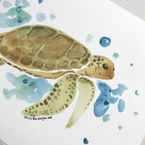 May include: Watercolour painting of a sea turtle swimming in blue water with green and blue watercolour splatter. The turtle is brown with a green shell and has a light brown face. The painting is signed "Suzi Blue Designs 2018".
