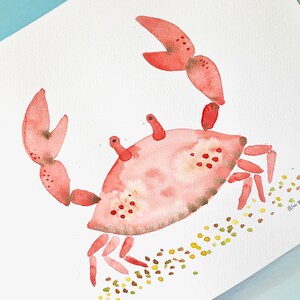 May include: Watercolor painting of a pink crab with yellow and brown dots on a white background. The crab is walking to the right and has its claws raised. The artist's signature is in the bottom right corner.