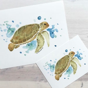 May include: Two watercolour paintings of sea turtles swimming in the ocean. The larger painting shows a turtle swimming to the right, while the smaller painting shows a turtle swimming to the left. Both paintings feature blue and green watercolour washes to represent the ocean.