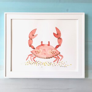May include: A watercolor painting of a pink crab on a white background with a light brown sand texture. The crab is facing forward with its claws raised. The painting is framed in a white frame.