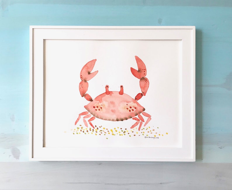 May include: A watercolor painting of a pink crab on a white background with a light brown sand texture. The crab is facing forward with its claws raised. The painting is framed in a white frame.