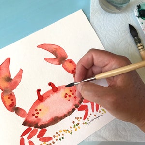 May include: A watercolor painting of a red crab with brown accents on a white background. The crab is surrounded by small yellow and green dots. The artist's signature is in the bottom right corner.