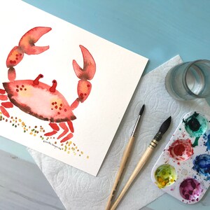 May include: A watercolor painting of a red crab on a white background with yellow and green dots representing sand. The painting is on a white sheet of paper with a glass of water, a paint palette, and two paintbrushes on a blue surface.