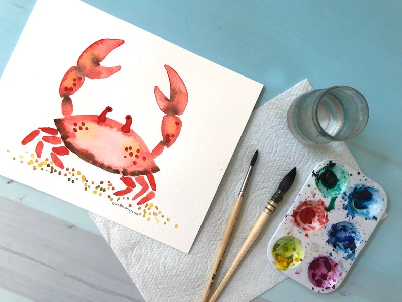 May include: A watercolor painting of a red crab on a white background with yellow and green dots representing sand. The painting is on a white sheet of paper with a glass of water, a paint palette, and two paintbrushes on a blue surface.