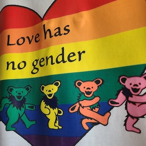 Puede incluir: Camiseta blanca con un diseño de arcoíris en forma de corazón y el texto "Love has no gender". Debajo, cuatro osos bailando en verde, amarillo, naranja y rosa.