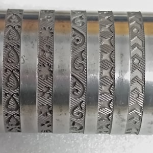 Könnte beinhalten: Eine Metallrolle mit sechs verschiedenen Prägemustern. Die Muster umfassen Herzen, Wirbel, Sterne und geometrische Designs.