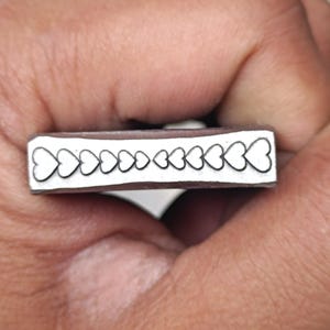 May include: A silver-coloured rectangular metal object featuring a series of heart-shaped cutouts. The hearts vary in size, creating a decorative pattern. The object is held in a hand.