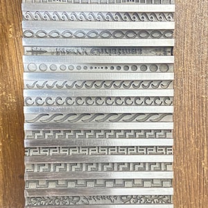 May include: A metal plate featuring a range of engraved designs, including geometric patterns, lines, and swirls. The plate is silver in colour and has a smooth, polished surface.