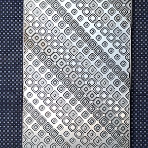 May include: A rectangular metal plate with a repeating pattern of squares and circles. The plate has a textured surface with a silver finish. The background is a dark blue fabric with a small checkered pattern.
