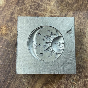 May include: A silver metal stamp with a design of a crescent moon and sun with a face, surrounded by stars.
