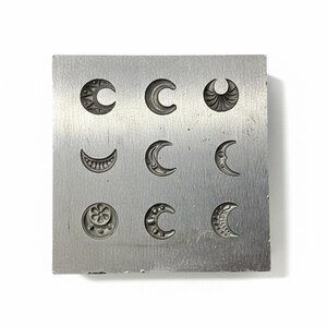 Könnte beinhalten: Ein quadratischer Stempelblock aus silberfarbenem Metall mit neun verschiedenen Mondsichel-Designs. Die Monde variieren im Stil, einige weisen detaillierte Muster auf, andere sind einfache Kurven. Der Block ist vor einem weißen Hintergrund platziert.
