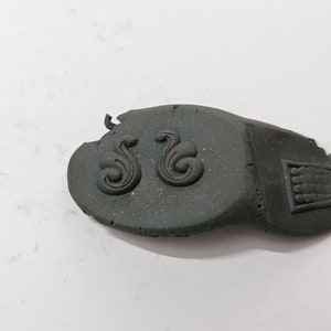 May include: A close-up of a dark gray, textured, rectangular piece of clay with two decorative swirls and a small rectangular design.
