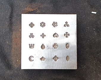 May include: A square metal stamp with various small decorative shapes, including flowers, hearts, and crosses. The stamp is made of a silver-colored metal and is designed for creating detailed impressions. The shapes are evenly spaced across the surface.