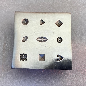 Könnte beinhalten: Ein silberner Metallstempel für Schmuck mit neun verschiedenen eingravierten Designs. Die Designs umfassen einen Kreis, ein Dreieck, eine Raute, ein Blatt, ein Oval, eine Spirale, eine Blume, ein Quadrat und ein Herz.