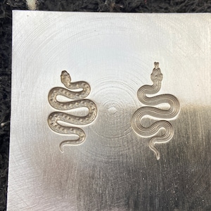 May include: Two snake designs are engraved into a silver metal plate. The snakes are facing to the right and are in a coiled position.