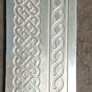 Puede incluir: Primer plano de una placa de metal plateado con dos patrones en relieve diferentes. El patrón de la izquierda es un diseño complejo e entrelazado, mientras que el patrón de la derecha es un diseño simple de eslabones de cadena.