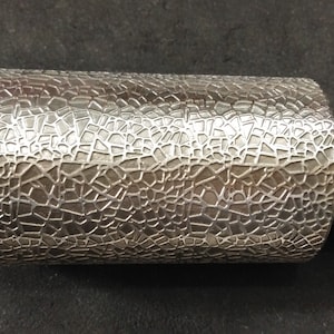 Könnte beinhalten: Ein zylindrisches Metallwerkzeug mit strukturierter Oberfläche, das ein Muster unregelmäßiger Formen aufweist. Das Werkzeug hat an jedem Ende einen soliden, polierten Metallschaft. Der strukturierte Teil hat einen Durchmesser von etwa 7,6 cm.
