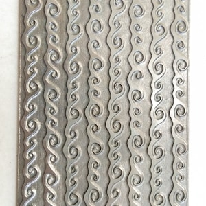 Op de afbeelding: Een zilveren metalen plaat met een herhalend patroon van golvende lijnen en swirls.