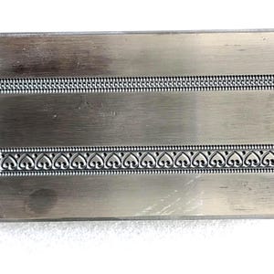 May include: A rectangular metal object with a brushed finish. It features two decorative borders: one with a dotted pattern and the other with a heart-shaped design. This could be a decorative element or part of a larger item.
