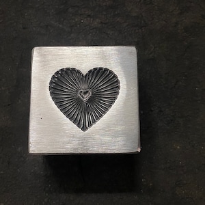 May include: A silver metal heart-shaped mold with a detailed design. The mold is square and has a smooth surface.