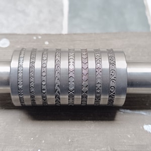Puede incluir: Un rodillo de metal plateado con varios diseños grabados. El rodillo se utiliza para crear patrones en arcilla u otros materiales.