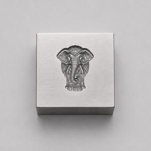 Könnte beinhalten: Ein quadratischer Block aus gebürstetem silberfarbenem Metall mit einem mittig geprägten Elefantenkopf-Design. Der Elefant ist mit detaillierten Ohren, Stoßzähnen und einem Rüssel dargestellt, in einem metallischen Finish.