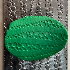 Op de afbeelding: Een ovaalvormig stuk klei in de kleur groen met een textuur van kleine cirkels. De klei is tegen een zilveren metalen oppervlak gedrukt met een vergelijkbaar patroon van cirkels.